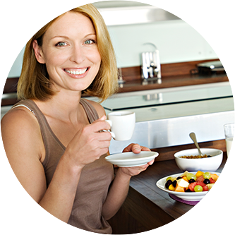 Smiling middle-aged blonde woman holding china cup and saucer, sitting in kitchen with fruit and cereal on table