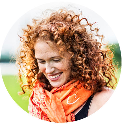 Headshot of smiling woman outdoors, with red curly hair and orange bandanna around neck